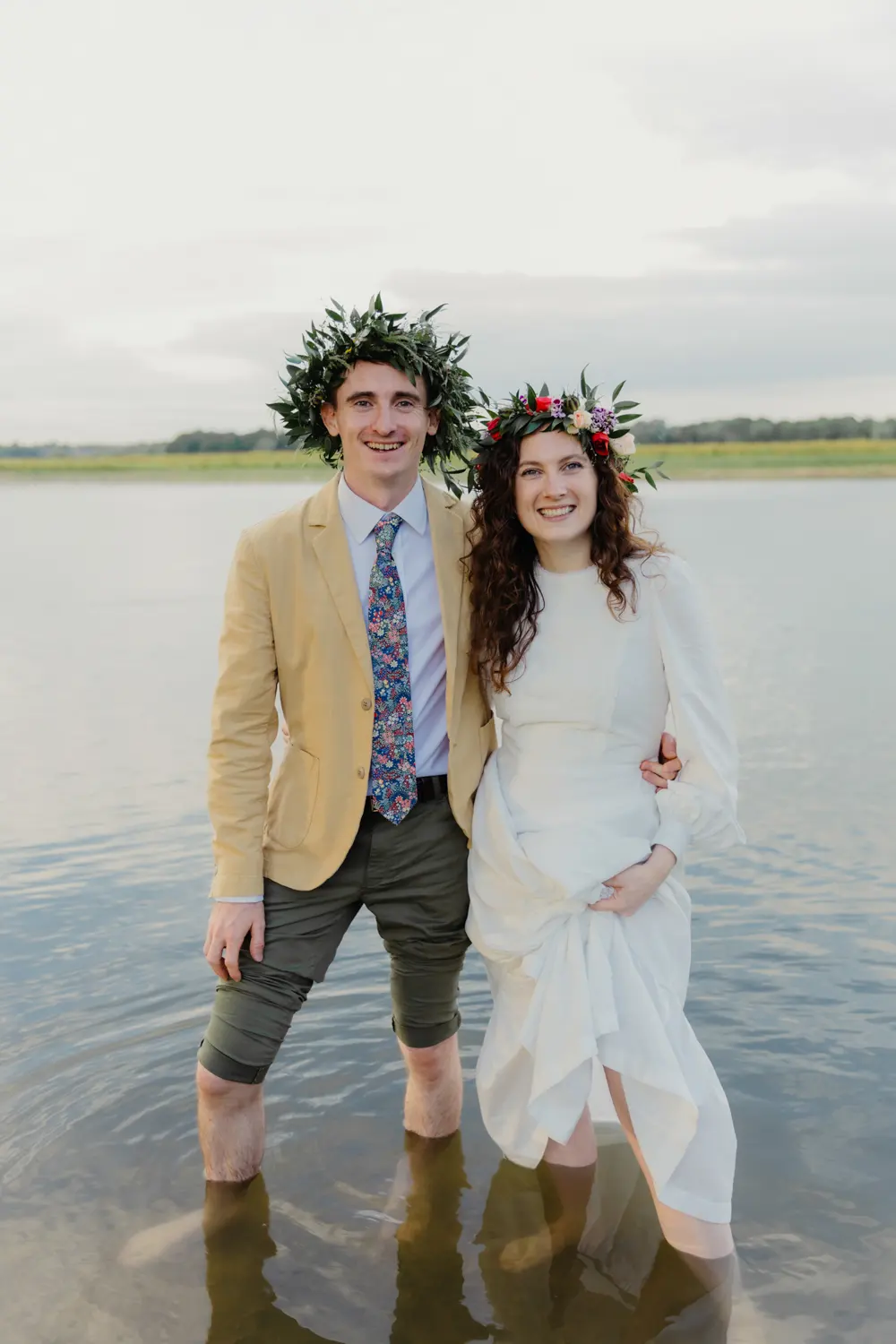 Bride and groom in flower and leaf crowns standing knee-deep in the water at Port Meadow
