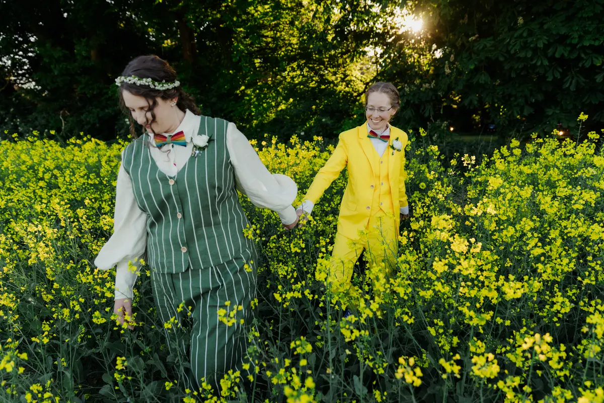 Two brides, one in a green striped suit and one in a yellow suit, walking hand in hand through a rapeseed field